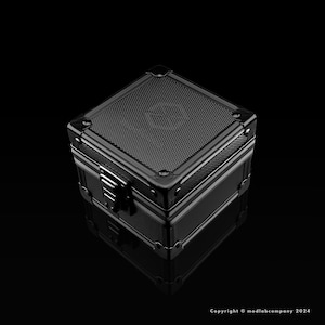 May include: A black, metallic box with a textured top and a silver clasp. The box has reinforced corners and a reflective surface, suggesting a high-quality construction. The box is sitting on a black surface.
