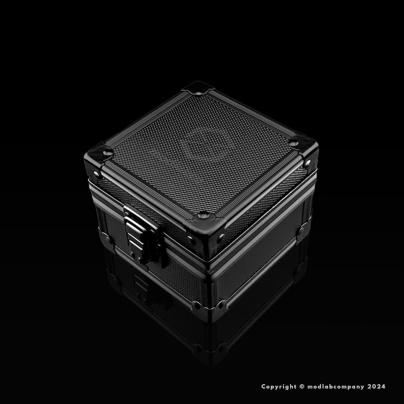 May include: A black, metallic box with a textured top and a silver clasp. The box has reinforced corners and a reflective surface, suggesting a high-quality construction. The box is sitting on a black surface.