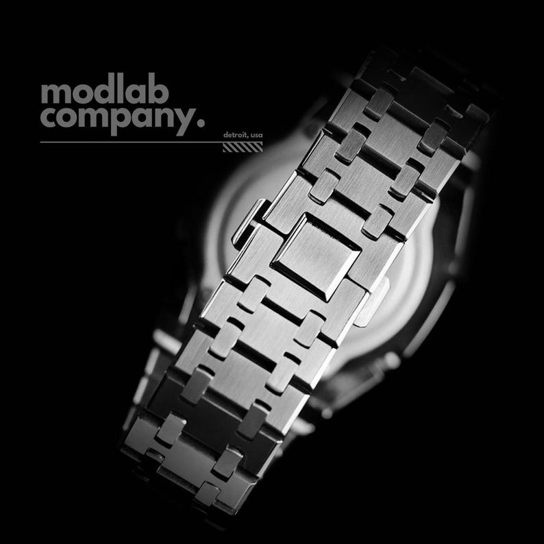 May include: Close-up of a stainless steel watch band with a geometric design. The watch band is silver and has a brushed metal finish. The watch face is visible in the background. The text "modlab company. detroit, usa" is in the upper left.