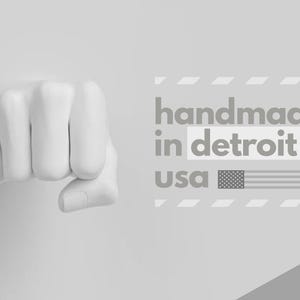 May include: A grayscale image featuring a clenched fist sculpture. The text "handmade in detroit usa" is displayed with an American flag graphic. The overall design is minimalist and modern, with a focus on the fist.