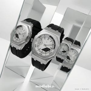 May include: Two silver watches with black bands are displayed in front of a mirrored surface. The watches have white faces with black numbers and hands. The watches are angled towards the viewer, showcasing their design. The image is well-lit, highlighting the watches' details.