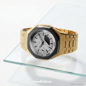 Op de afbeelding: Een goudkleurig polshorloge met een zwarte lunette en een zilveren wijzerplaat. Het horloge heeft een rechthoekig ontwerp met een gouden band. De wijzerplaat geeft de tijd en andere informatie weer. Het horloge wordt gepresenteerd op een glazen oppervlak.