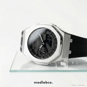 Op de afbeelding: Een zilverkleurig G-Shock Casio horloge met een zwarte wijzerplaat en zwarte band. Het horloge toont de tijd als 10:10. Het horloge ligt op een helder glazen oppervlak.