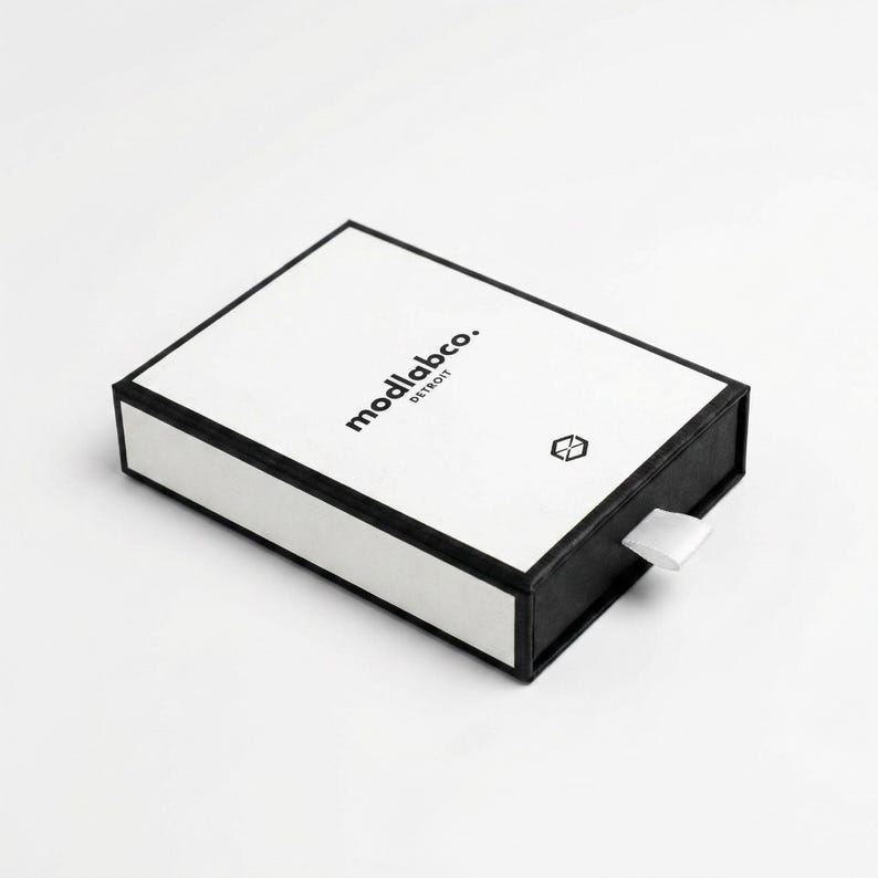 Puede incluir: Caja rectangular en blanco y negro con el texto "modlabco. DETROIT" y un peque&ntilde;o s&iacute;mbolo geom&eacute;trico. La caja tiene una tapa blanca y una base negra, con un tirador de cinta blanca.
