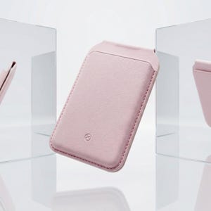 Puede incluir: Un tarjetero de tel&eacute;fono de cuero rosa claro con un borde cosido y un peque&ntilde;o logotipo. El tarjetero tiene un bolsillo en la parte delantera y una parte superior curvada. El fondo es una superficie reflectante.