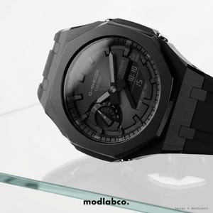 Op de afbeelding: Een close-up van een zwarte G-Shock Casio horloge. Het horloge heeft een digitale display met de tijd, met een zwarte band en een getextureerde bezel. Het horloge wordt getoond op een helder oppervlak.