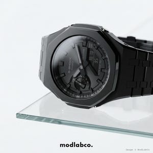 May include: A black G-Shock Casio watch with a dark grey face, showing the time 10:10. The watch has a square case and a linked strap. The watch is displayed on a glass surface.
