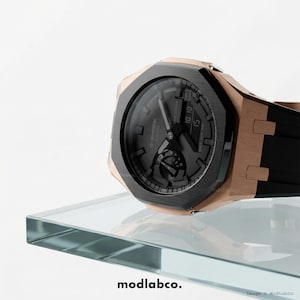 Op de afbeelding: Een G-Shock Casio horloge met een zwarte wijzerplaat en roségouden accenten. Het horloge heeft een zwarte band en wordt gepresenteerd op een glazen plank. De tijd die wordt weergegeven is 10:10. De merknaam "modlabco." is onderaan zichtbaar.