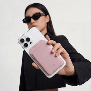 Puede incluir: Un tel&eacute;fono inteligente blanco con un tarjetero de cuero rosa adjunto. El tarjetero tiene dos ranuras y un borde cosido. El tel&eacute;fono tiene una c&aacute;mara de triple lente y se sostiene contra una blazer negra.