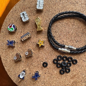 May include: A black braided leather bracelet with a silver clasp, accompanied by various metal charms including FIFA logos, a gold trophy, a star, a rocket, and Texas-shaped icons, alongside a small collection of black rubber O-ring spacers.