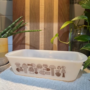 Puede incluir: Un molde rectangular para pan de vidrio lechoso blanco con un patrón gráfico repetitivo de color marrón que muestra diversas formas de hongos a lo largo de los lados exteriores. El recipiente tiene un acabado liso y brillante con asas.