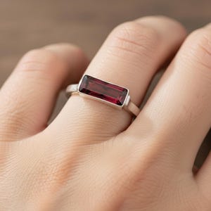 Op de afbeelding: Een zilveren ring met een rechthoekige, dieprode edelsteen. De edelsteen is gezet in een bezel setting en de band is van gepolijst zilver. De ring wordt op een vinger getoond, waarbij het ontwerp en de kleur worden benadrukt.