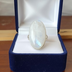 Op de afbeelding: Zilveren ring met een grote, ovale maansteen, gepresenteerd in een blauwe fluwelen doos. De maansteen is melkachtig wit met blauwe iriserende tinten. Een opvallend sieraad.