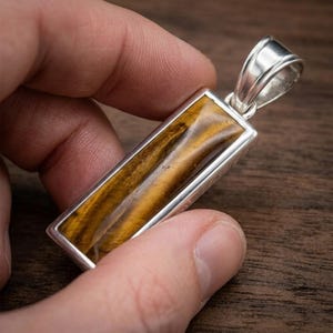 Puede incluir: Un colgante rectangular con una piedra de ojo de tigre pulida engastada en un marco plateado. El colgante tiene una argolla para una cadena. La piedra ojo de tigre muestra bandas de marrón dorado y amarillo.