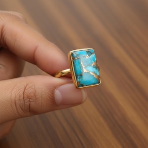 May include: A gold-colored ring featuring a rectangular turquoise stone with brown veining. The ring has a simple band and a bezel setting that frames the stone. The turquoise stone has a polished finish.