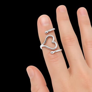 May include: A silver-colored ring with a heart-shaped design. The ring wraps around the finger with a heart in the center and two curved bars with rounded ends. The ring is shown on a finger against a black background.