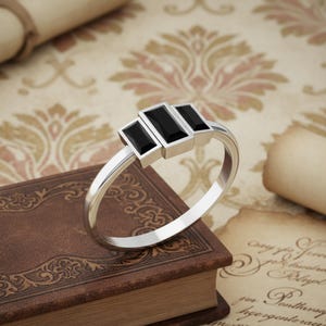 May include: A silver ring with three rectangular black gemstones set in a row. The ring is displayed on a vintage book with a decorative cover, against a backdrop of aged paper and floral patterns.