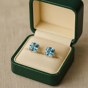 May include: A pair of square, light blue gemstone stud earrings set in silver, displayed inside an open, dark green jewelry box with a cream-colored interior. The earrings are the focal point, showcasing their vibrant color and design.