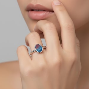 Può includere: Un anello d'argento con una gemma ovale che mostra una sfumatura di blu e rosso. L'anello è indossato su un dito, evidenziando la fascia in argento lucido e i colori vivaci della pietra. Il design è semplice ma elegante.