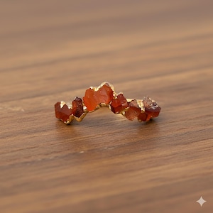 May include: A gold ring adorned with raw, orange-red gemstones. The stones are arranged along the band, forming an irregular, natural shape. The ring is presented on a wooden surface, highlighting its earthy tones and unique design.