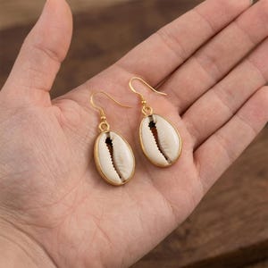 Puede incluir: Un par de pendientes colgantes con conchas de cauri blancas naturales engastadas en metal dorado. Cada concha cuelga de un sencillo gancho dorado, mostrando la textura orgánica y la ranura central característica de la concha.