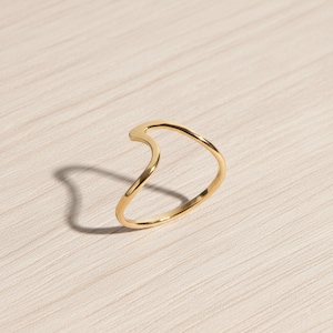 May include: A gold-colored ring with a curved, wave-like design. The ring is set against a light-colored wooden surface, with a shadow cast to the left. The ring's smooth, polished surface reflects the light.