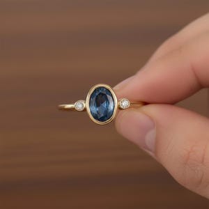 Könnte beinhalten: Ein goldener Ring mit einem ovalen, tiefblauen Edelstein als Mittelpunkt, flankiert von zwei kleineren, runden, klaren Edelsteinen. Der Ring wird vor einem verschwommenen Hintergrund gehalten, der das elegante Design hervorhebt.