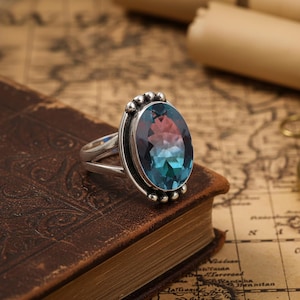 Può includere: Anello in argento con una gemma ovale cangiante, che passa dal verde-blu al rosso-viola. L'anello, con gambo diviso, è appoggiato su un libro rilegato in pelle marrone.