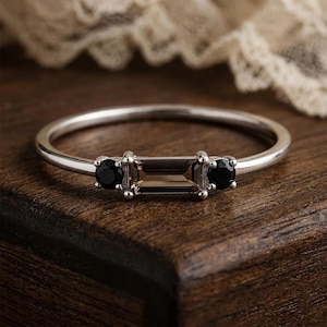 Puede incluir: Un anillo de plata con una piedra central rectangular de color ahumado, flanqueada por dos piedras negras redondas. El anillo tiene una banda delgada, que muestra un dise&ntilde;o minimalista. Las piedras preciosas est&aacute;n engastadas con garras.