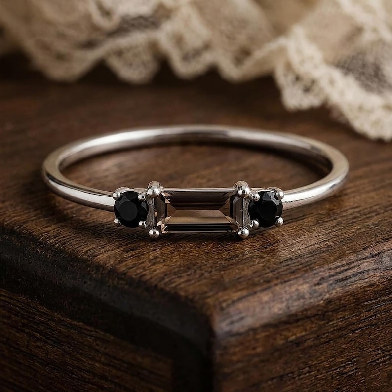 Puede incluir: Un anillo de plata con una piedra central rectangular de color ahumado, flanqueada por dos piedras negras redondas. El anillo tiene una banda delgada, que muestra un dise&ntilde;o minimalista. Las piedras preciosas est&aacute;n engastadas con garras.
