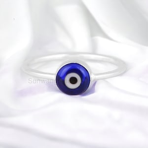 Può includere: Un anello d'argento con un ciondolo a forma di occhio turco blu. L'anello è su uno sfondo bianco.