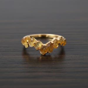 Könnte beinhalten: Ein goldfarbener Ring mit einer Reihe unregelmäßig geformter, hellgelb-oranger Edelsteine. Die Edelsteine sind zu einem einzigartigen, organischen Design zusammengefasst. Der Ring liegt auf einer dunklen Holzoberfläche.