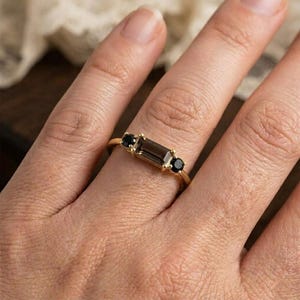 Puede incluir: Un anillo de oro con una gema rectangular de color marr&oacute;n ahumado flanqueada por dos gemas negras redondas m&aacute;s peque&ntilde;as. El anillo est&aacute; engarzado en una delgada banda de oro, mostrando un dise&ntilde;o minimalista.