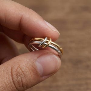 Może przedstawiać: Zbliżenie pierścionka z motywem węzła. Pierścionek posiada złoty pasek przeplatany dwoma srebrnymi paskami. Paski są gładkie i błyszczące, tworząc kontrast między złotymi i srebrnymi odcieniami. Pierścionek jest trzymany między palcami.