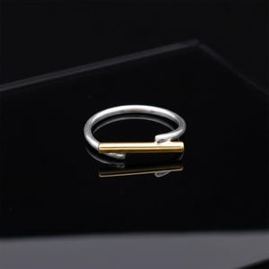 May include: A minimalist ring featuring a silver band and a gold-coloured horizontal bar. The ring's design is simple and modern, with a sleek, polished finish. The ring is set against a black background.