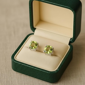 May include: A pair of square-cut, light green peridot stud earrings in a dark green jewelry box. The earrings are set in silver and are displayed on a cream-coloured interior. The box is open, showcasing the earrings.