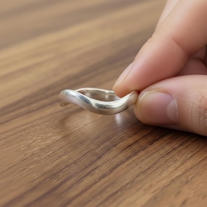 May include: A silver ring with a wavy design. The ring is held between fingers, showcasing its smooth, polished surface. The ring's unique shape and metallic sheen are highlighted against a wooden background.