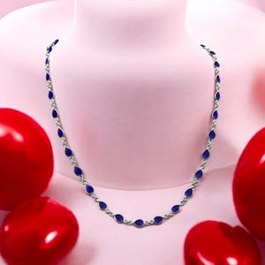 May include: A silver necklace with a delicate chain and blue teardrop-shaped charms. The necklace is displayed against a pink background.