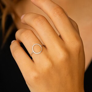 Op de afbeelding: Een zilveren ring met een minimalistisch ontwerp. De ring heeft een dunne band en een cirkelvormige, open vorm als middelpunt. De ring wordt getoond aan de vinger van een persoon, met de hand in focus tegen een vage achtergrond.