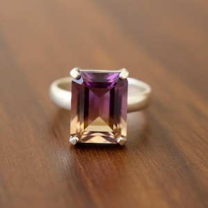 May include: A silver ring with a large, rectangular gemstone displaying a colour gradient. The stone transitions from deep purple to golden-yellow. The ring is set on a wooden surface.