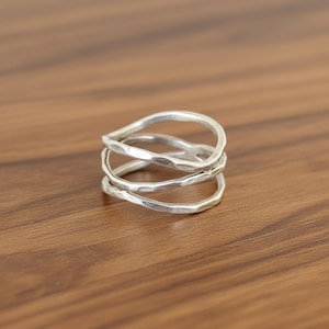 Könnte beinhalten: Ein silberner Ring mit einem einzigartigen, geschichteten Design. Der Ring hat drei ineinander verschlungene Bänder mit einer gehämmerten Textur, die einen modernen und künstlerischen Look erzeugt. Der Ring wird auf einer Holzoberfläche präsentiert.