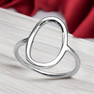 May include: A silver ring with an open oval design. The ring is made of polished metal and has a smooth, reflective surface. The oval shape is the focal point, creating a modern and minimalist aesthetic. The ring sits on a light-colored wooden surface.