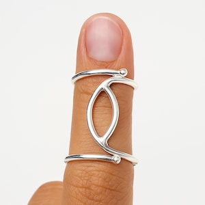 May include: A silver ring with an open, geometric design. The ring features a central oval shape and curved lines that wrap around the finger. The ring is worn on a finger, showcasing its modern and minimalist style.