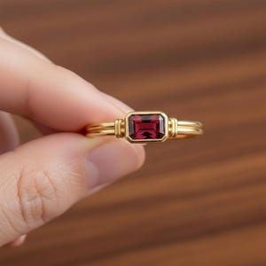Peut inclure: Une bague en or avec une pierre précieuse rectangulaire rouge foncé. La bague a un double anneau et des éléments décoratifs sur les côtés de la pierre précieuse. La bague est tenue par les doigts d'une personne.