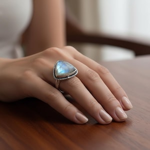 Könnte beinhalten: Ein silberner Ring mit einem großen, dreieckigen, schillernden Mondstein. Der Mondstein hat eine blau-weiße Farbe und ist in eine gedrehte, seilartige Lünette gefasst. Der Ring befindet sich an einer Hand mit neutraler Maniküre.