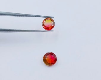 May include: Two round, faceted gemstones with a red, orange, and yellow gradient. The gemstones are held by tweezers.