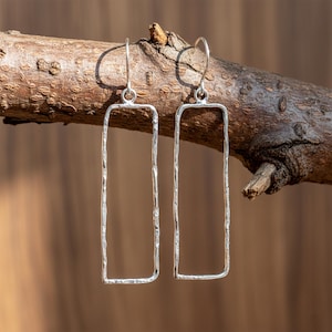 Op de afbeelding: Een paar zilveren rechthoekige oorhangers met een gehamerde textuur. De oorbellen hangen aan gebogen haken en worden gepresenteerd tegen een vage bruine achtergrond, op een tak.