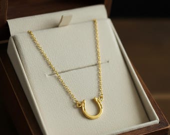 May include: A gold-colored horseshoe pendant necklace displayed in a wooden jewelry box. The necklace features a delicate chain and a horseshoe charm, symbolizing luck and fortune. The box has a cream-colored interior.