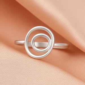 May include: A silver ring with a spiral design. The ring has a thin band and a circular design with two concentric circles. The ring is set against a peach-coloured fabric background.