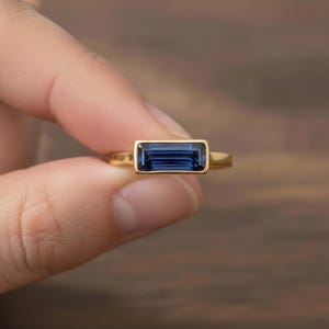 May include: A gold ring with a rectangular, deep blue gemstone. The gemstone is set in a bezel setting, and the ring band is a simple, polished gold. The ring is held against a blurred wooden background.